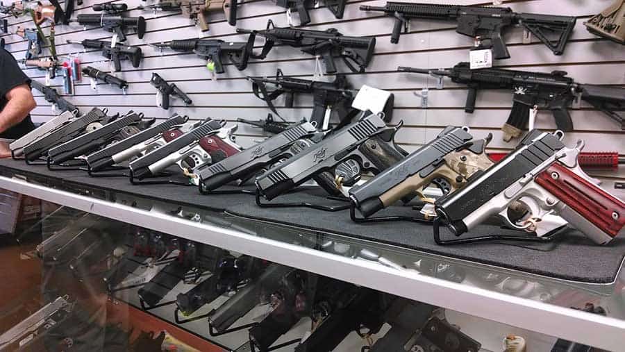 A gun store display features a variety of pistols lined up on a counter, with rifles mounted on the wall behind them. Each firearm is securely positioned, showcasing different styles and designs.