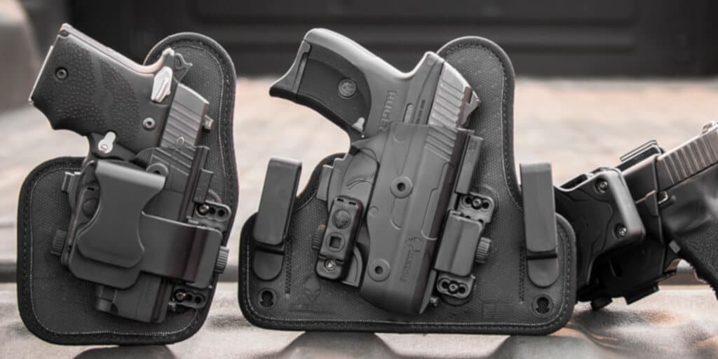 Several holstered handguns are displayed on a surface. The holsters are black with attached clips, designed for secure carrying. The background is blurred, emphasizing the firearms in the foreground.