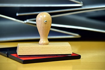 A wooden rubber stamp resting on a red ink pad placed on a wooden surface, with several black binders stacked in the background.