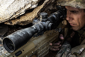 A soldier in camouflage holds a sniper rifle, partially concealed by rocky terrain. His focused gaze is through the rifle scope, suggesting readiness and alertness in a tactical position.