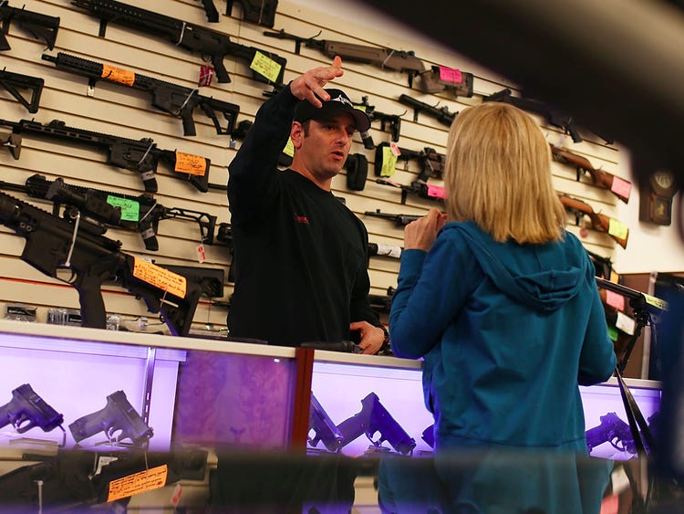 A man in a cap gestures while speaking to a woman in a blue hoodie across a glass counter in a gun store. Various rifles are displayed on the wall behind them with different colored price tags.