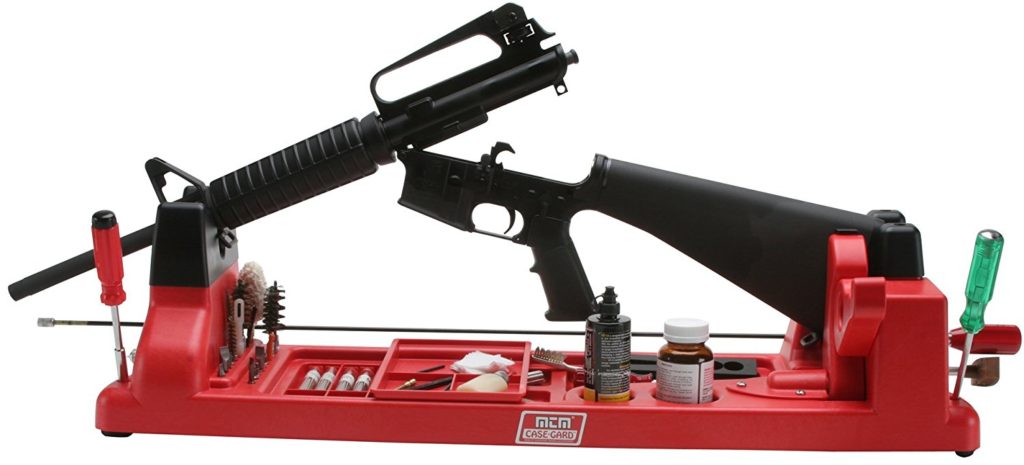 A rifle is securely placed on a red gun cleaning and maintenance stand. The stand has compartments holding various cleaning tools, brushes, and bottles of cleaning solutions.