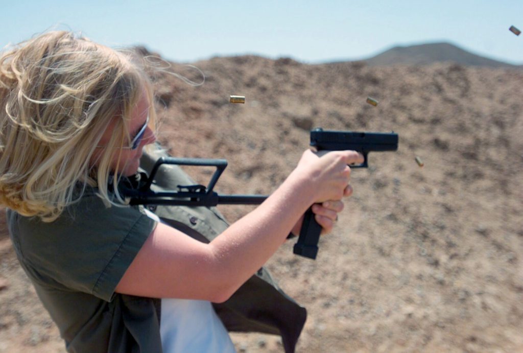 A person with light hair, wearing sunglasses and a green jacket, is shooting a handgun outdoors. Spent casings are ejected from the gun as they stand in front of a rocky, barren landscape.
