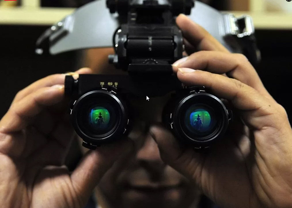 A person looking through night vision goggles, with a reflection of a figure visible in the lenses. The goggles have a metallic frame and bulky design. The scene is dimly lit, suggesting a focus on technical or surveillance activity.