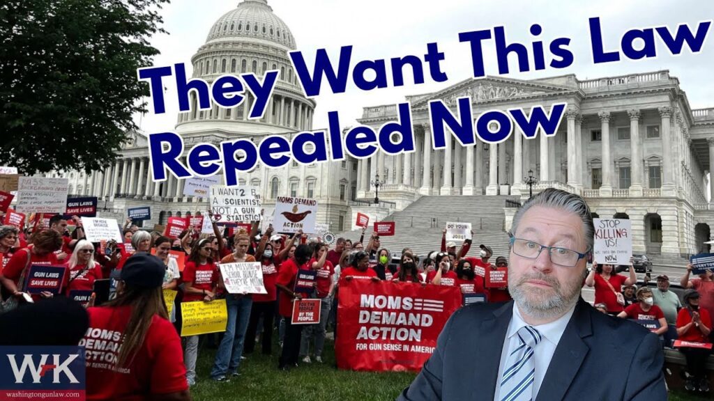 A large group of people hold up signs outside a government building with a dome, protesting for gun legislation change. A man in a suit is in the foreground. Text reads, "They Want This Law Repealed Now.