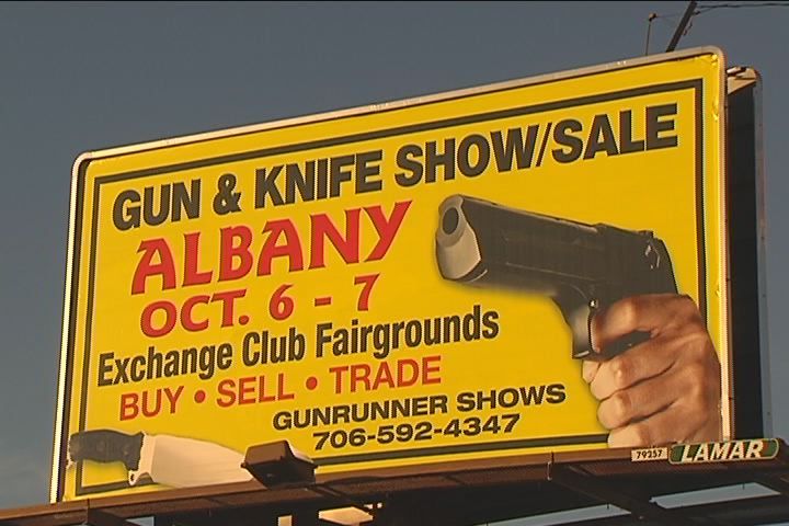 A billboard advertising a gun and knife show/sale in Albany on October 6-7, located at the Exchange Club Fairgrounds. It features a hand holding a pistol. Contact information and details about buying, selling, and trading are included.