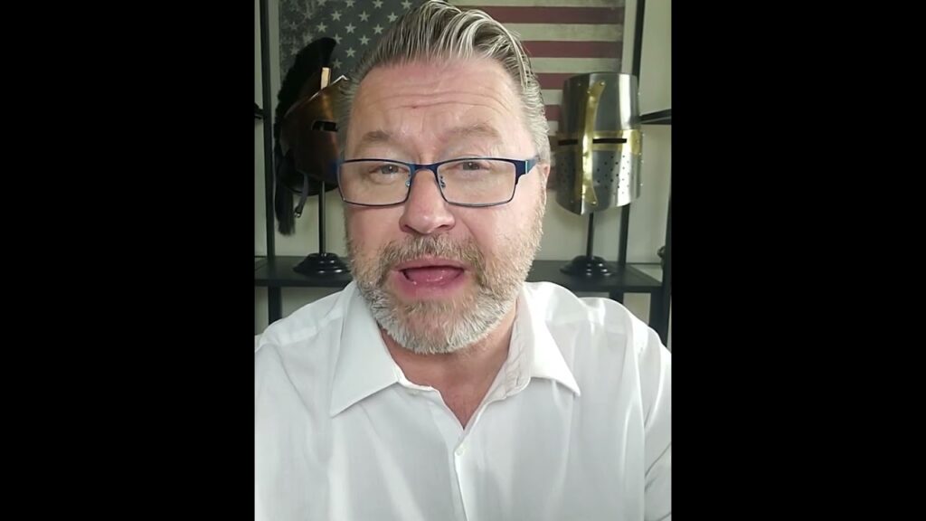 A man with glasses and a beard wearing a white shirt stands in front of a shelf displaying knight helmets. An American flag is partially visible in the background.