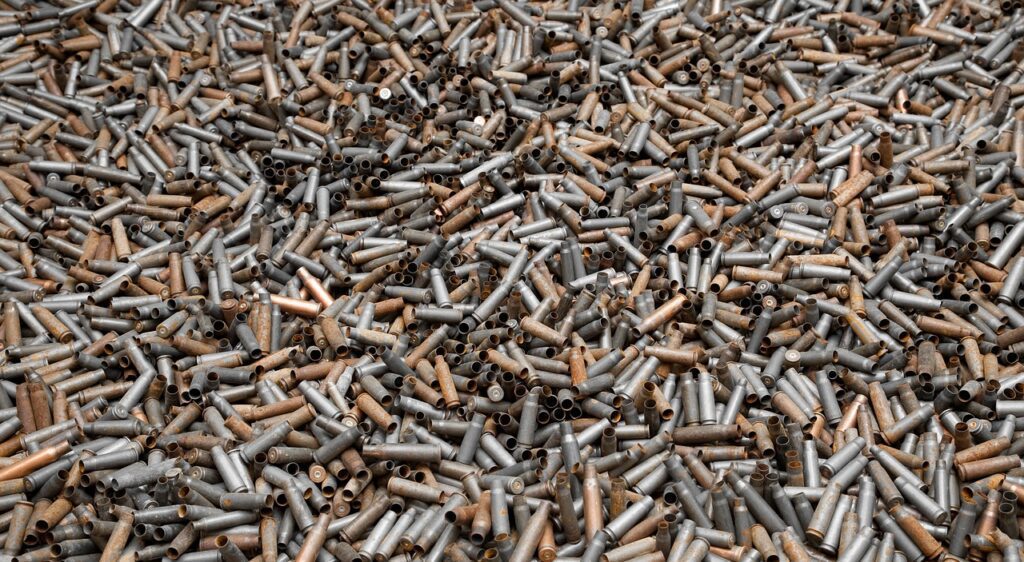 A large collection of empty bullet casings in various shades of silver and bronze are scattered randomly, filling the entire frame. Some casings are upright while others lie horizontally, creating a textured, metallic surface.