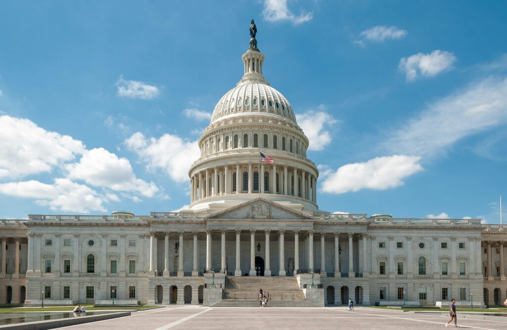 The image shows the United States Capitol building in Washington, D.C., under a clear blue sky. The dome is prominent, and an American flag waves in front. There are a few people walking near the entrance.