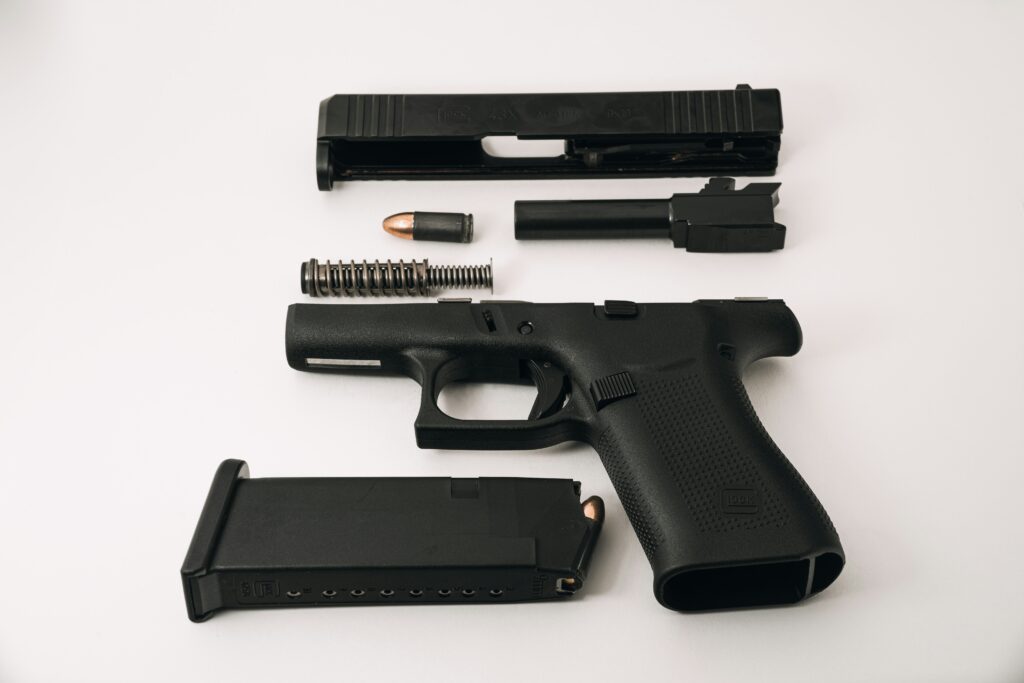 A disassembled black handgun laid out on a white surface, showing its components including the slide, barrel, recoil spring, frame, bullet, and magazine.