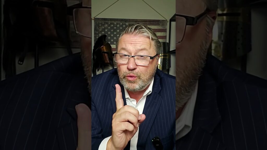 A man with glasses and a beard is wearing a pinstripe suit and gestures with his index finger raised. A flag and wooden decor are visible in the background.