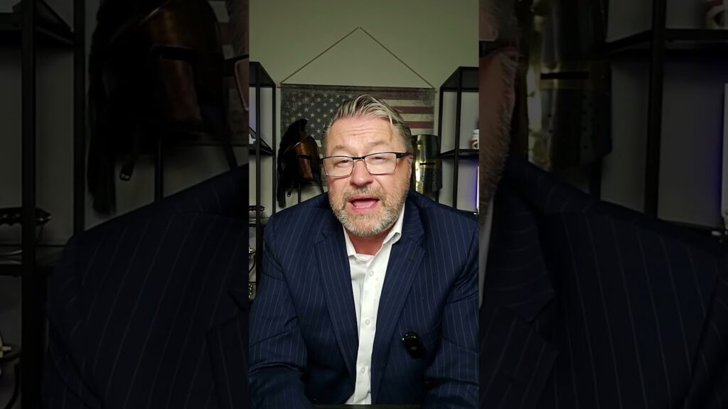 A man with glasses and a beard is speaking while wearing a navy pinstripe suit. He is indoors with an American flag in the background, and there are shelves with various objects behind him.