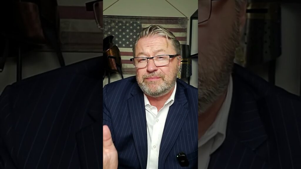 A man with glasses and a beard is speaking in front of an American flag backdrop. He is wearing a striped suit and a white shirt and appears to be gesturing with one hand. The image has a vertically cropped format.