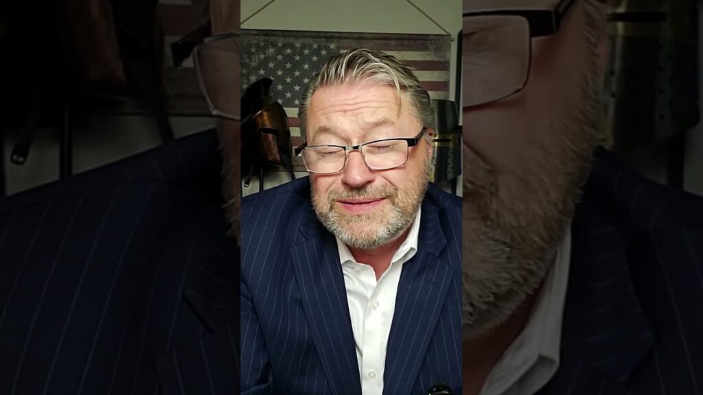 A man with glasses and a beard, wearing a pinstripe suit and white shirt, is talking. He is in a room with an American flag in the background.