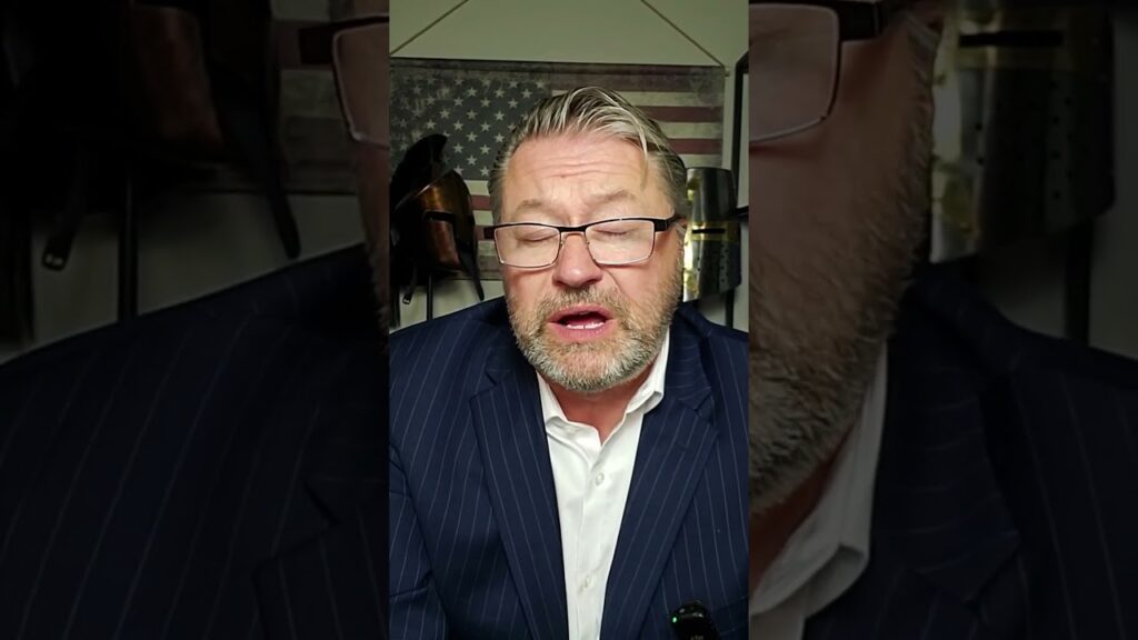 A bearded man in a suit and glasses is speaking. Behind him is an American flag and a wall with various objects, including a cowboy hat. The setting appears to be an indoor, well-lit room.