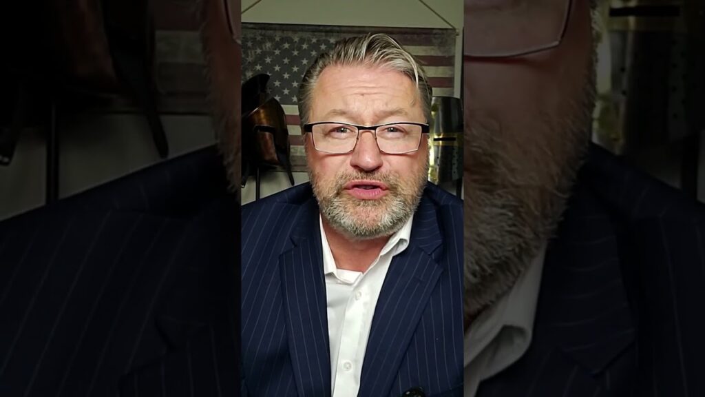 A middle-aged man with glasses and a beard, wearing a pinstriped suit, speaks to the camera with an American flag in the background.