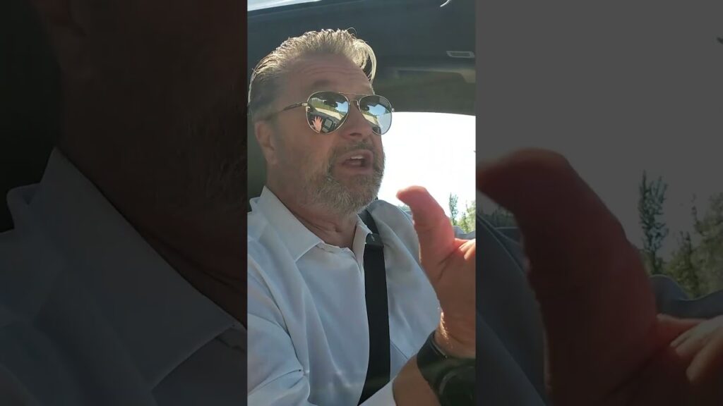 A man with sunglasses and a gray beard is driving a car. He wears a white shirt and gestures with his right hand. Sunlight reflects off his sunglasses.