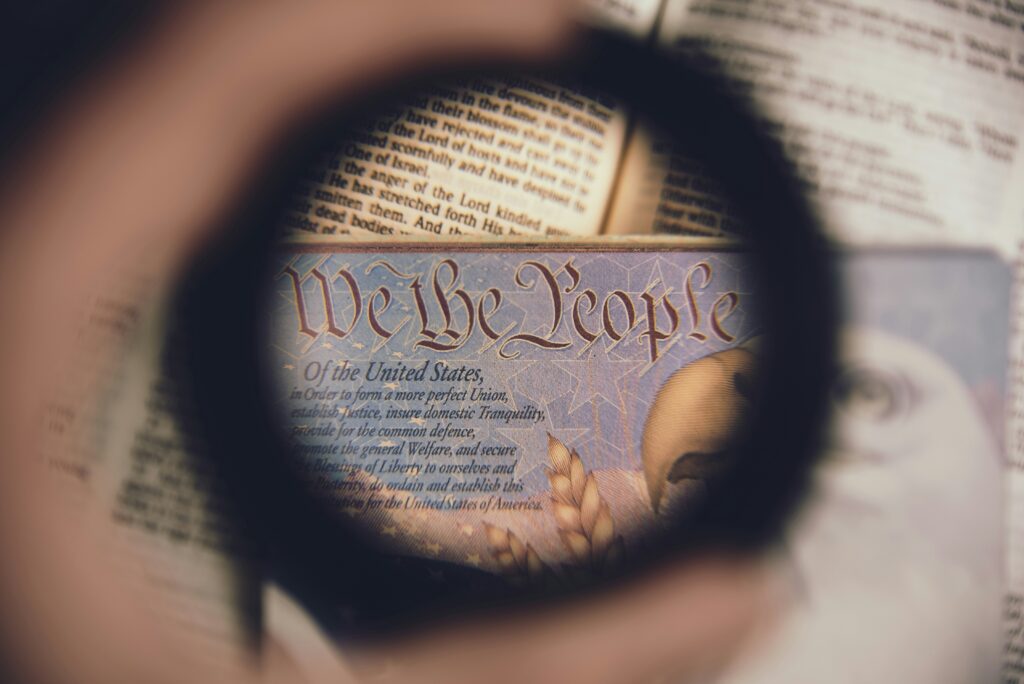 A magnifying glass focuses on the phrase "We the People" from the U.S. Constitution, with an American flag visible in the background. Text is partially obscured and appears on an open book beneath the lens.