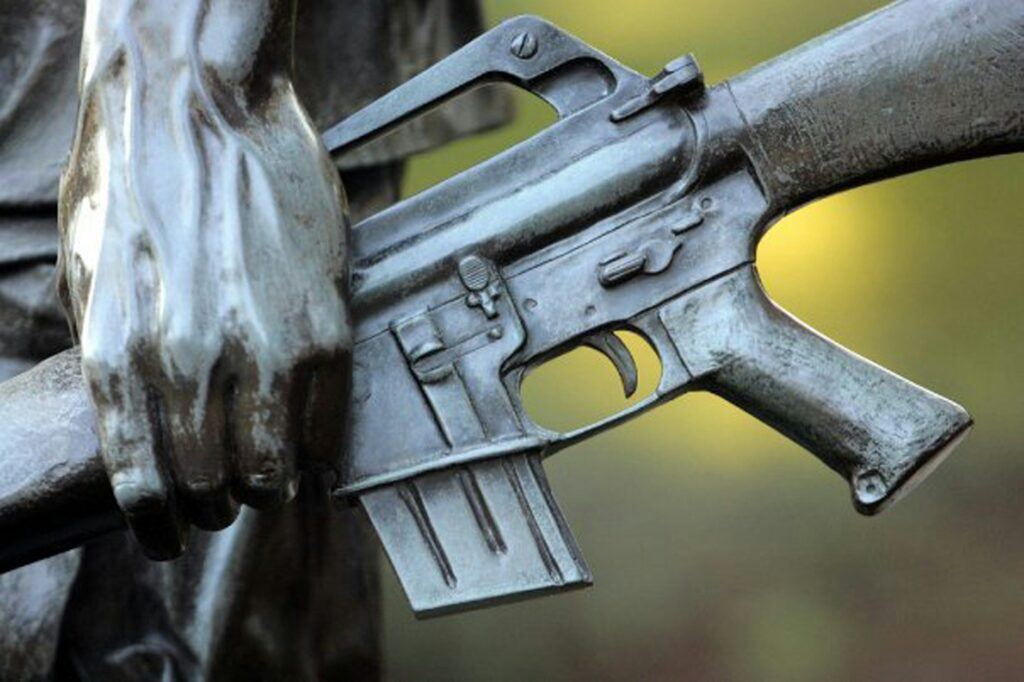Close-up of a statue depicting a person's hand holding a rifle. The statue is made of metal, with detailed features on both the hand and the weapon. The background is softly blurred with green and yellow hues.
