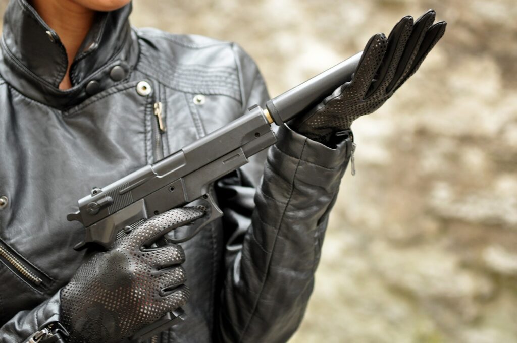 A person in a black leather jacket and gloves holds a silenced handgun. The background is a blurred, neutral color. The individual's face is partially visible, focusing on the firearm being examined.