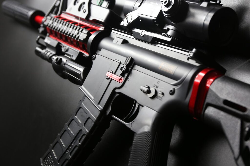 Close-up of a black and red tactical rifle on a dark surface. The image highlights the gun's detailed components, including the magazine, safety switch, and scope.