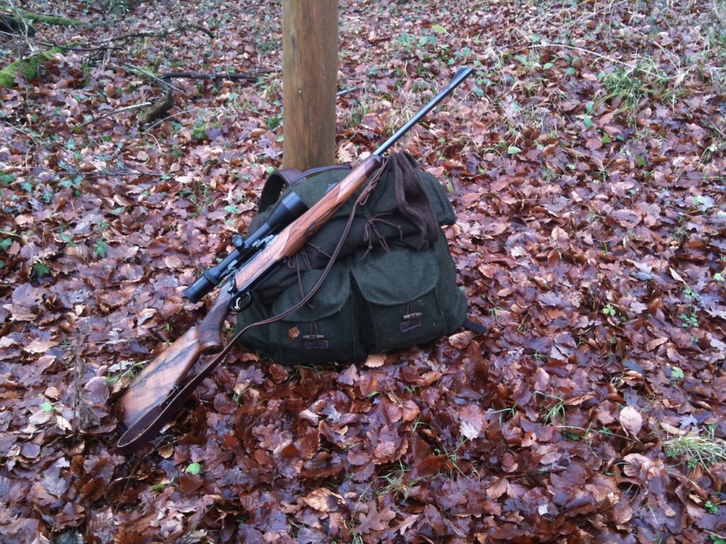 A rifle with a scope rests on top of a green backpack on the ground covered with brown leaves in a forest setting. A wooden post is visible in the background among bare branches.