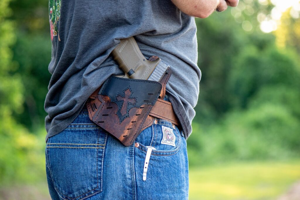 Person wearing jeans and a grey shirt with a holstered handgun on their hip. The holster has a decorative design. Greenery is visible in the background.