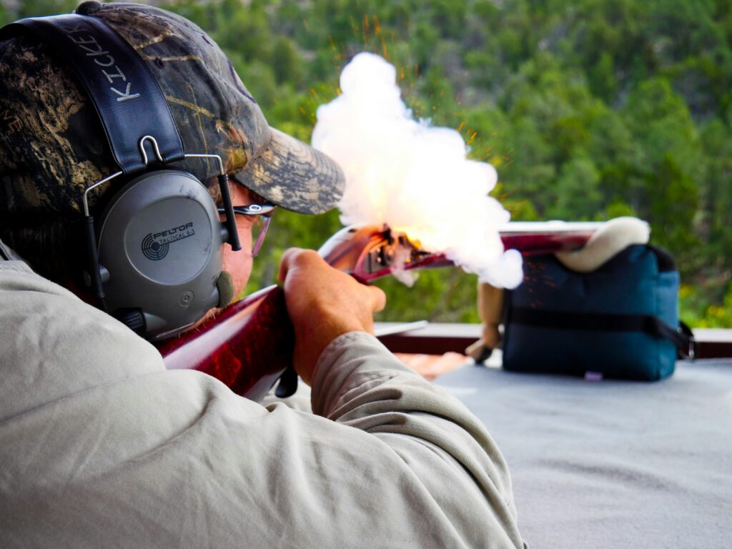 A person wearing a camouflage cap and ear protection is firing a muzzleloader rifle outdoors. White smoke and sparks are visible from the rifle's firing end. They are positioned at a shooting range with green trees in the background.