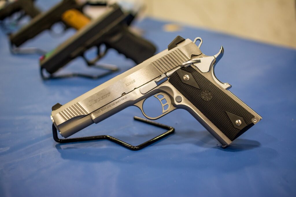 Close-up of a silver handgun with a black grip on a blue display stand. The gun is angled to the right, showing its side details, with other blurred firearms in the background.