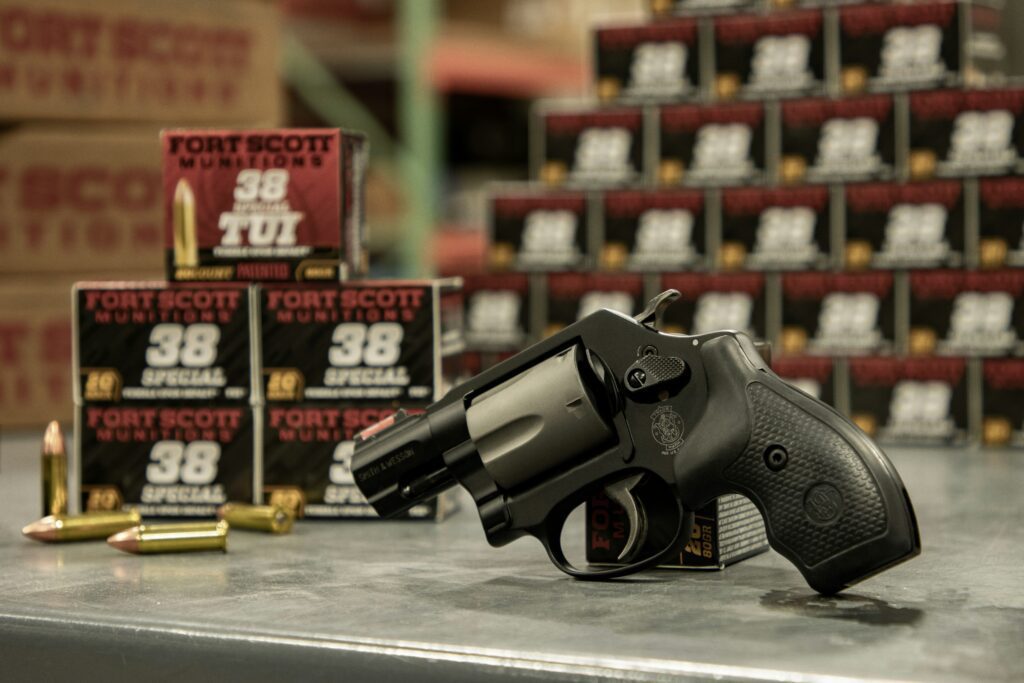 A black revolver is displayed on a counter with its side facing up, surrounded by assorted ammunition. In the background, there are numerous boxes labeled "Fort Scott Munitions .38 Special.