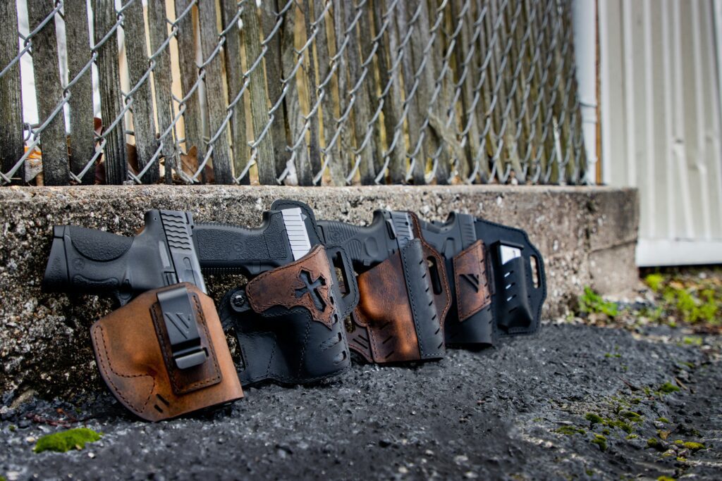 Several handguns in leather holsters are lined up on the ground in front of a chain-link fence. The setting appears to be outdoors, and the ground is asphalt with some scattered leaves and pebbles.