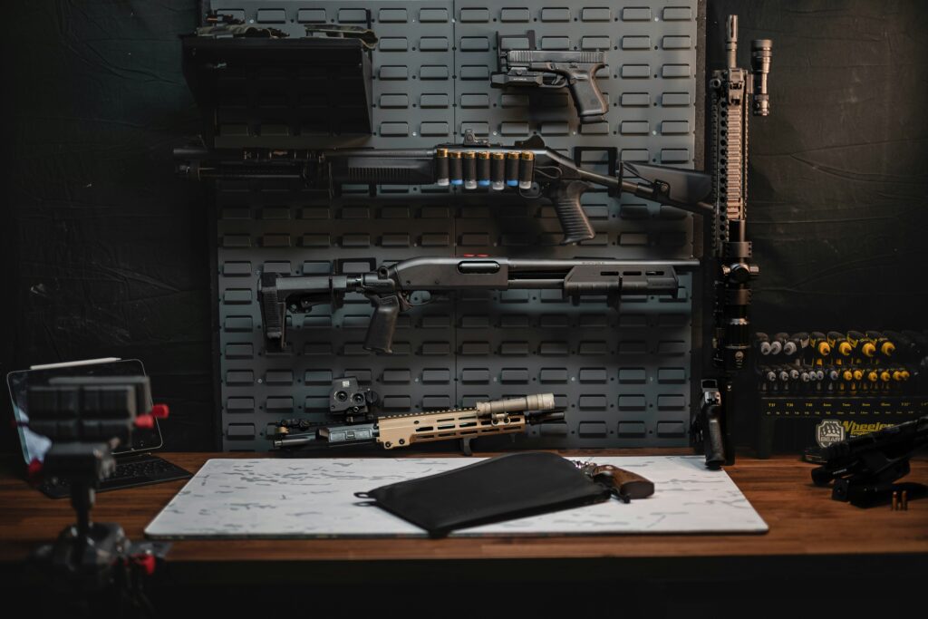 A workbench with various firearms mounted on a pegboard, including rifles and handguns. Ammunition is displayed on the right alongside other gun accessories. A laptop and black clipboard are placed on the wooden surface.