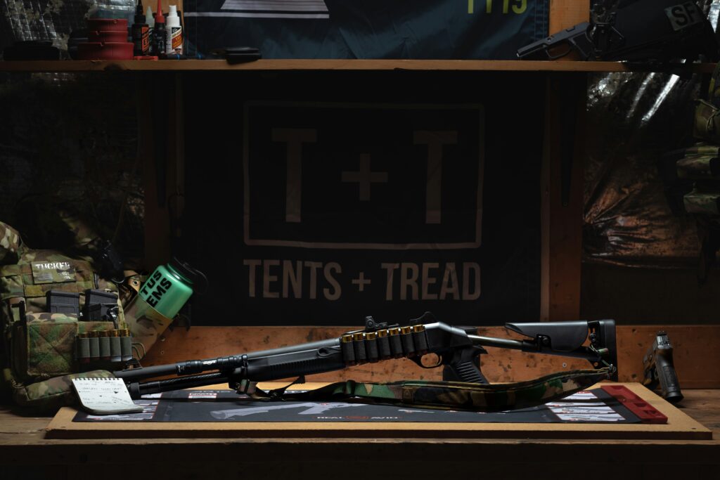 A rifle displayed on a wooden table with a camouflage strap and bullet pouch, surrounded by military gear and accessories. A "Tents + Tread" banner hangs in the background. The scene is dimly lit, highlighting the firearm and gear.