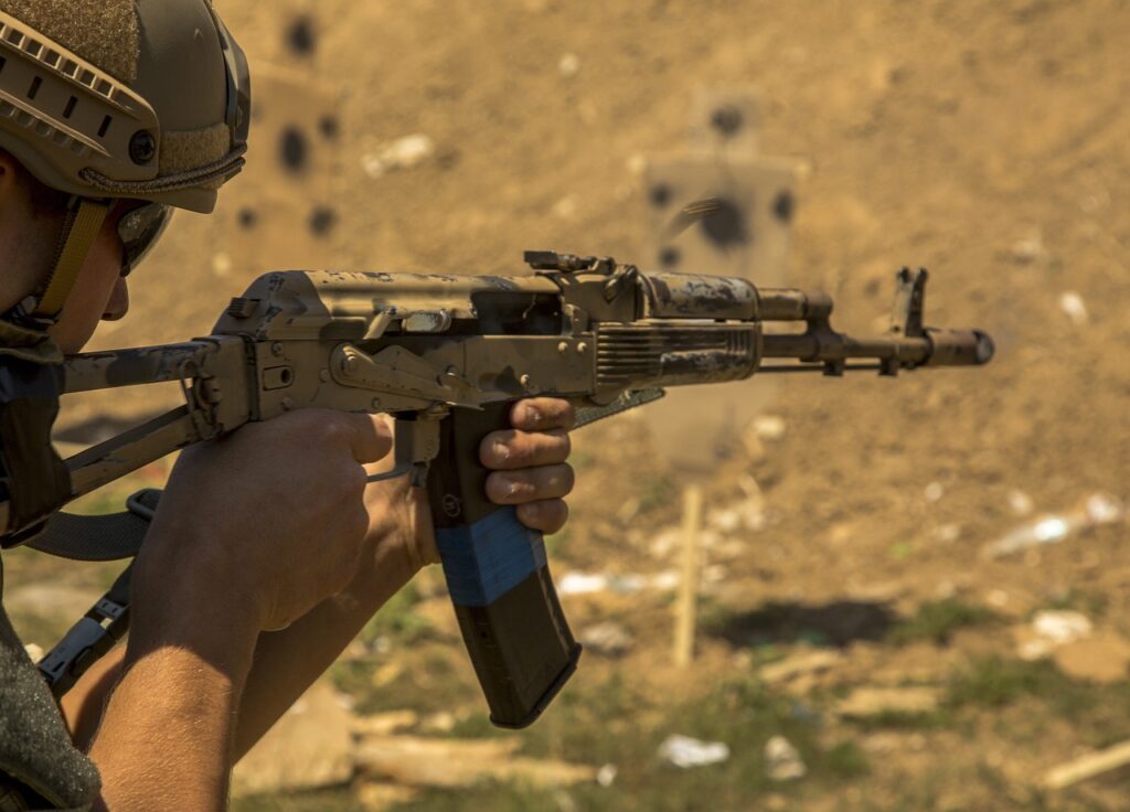 A person in tactical gear aims an assault rifle at a target during a shooting practice. The background is an outdoor shooting range with a dirt berm and paper targets.