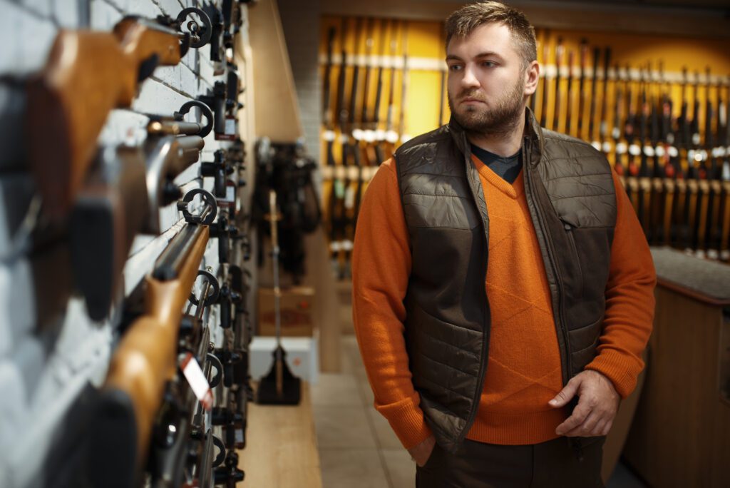 A man wearing an orange sweater and brown vest stands in a store, looking at a display wall filled with rifles. The background features rows of firearms on racks.