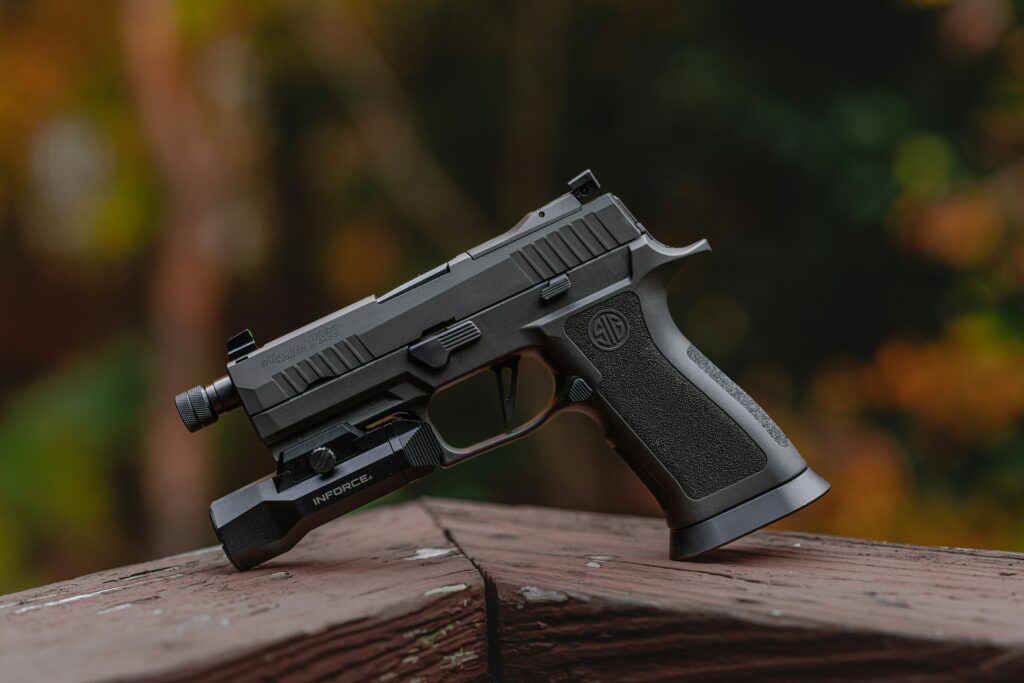 A black handgun with a tactical light attachment is resting on a wooden surface. The background is softly blurred with autumn foliage in shades of green, orange, and brown.