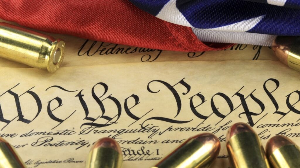 Close-up of the U.S. Constitution with the words "We the People" visible, surrounded by bullet casings. An American flag features prominently in the background, partially covering the document.