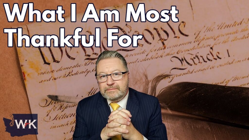 A man in a suit sits with his hands clasped, looking forward. Behind him is a text overlay that reads "What I Am Most Thankful For." The background features a close-up of a document with the words "We the People.