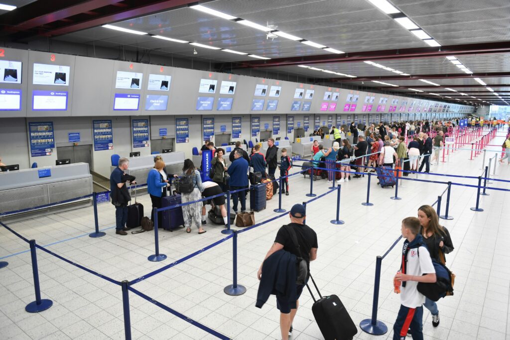 A busy airport check-in area with numerous people lining up at counters. There are digital screens above the counters. The floor is tiled, and stanchions with blue straps guide the lines. Passengers carry luggage, with some wearing casual attire.