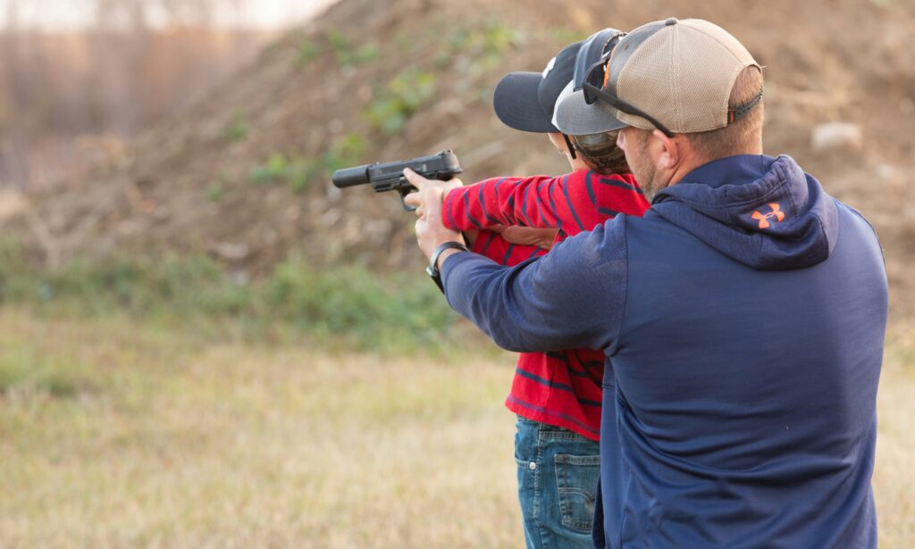 A man in a blue hoodie assists a child wearing a red striped shirt and a cap in aiming a gun outdoors, with a dirt mound in the background.