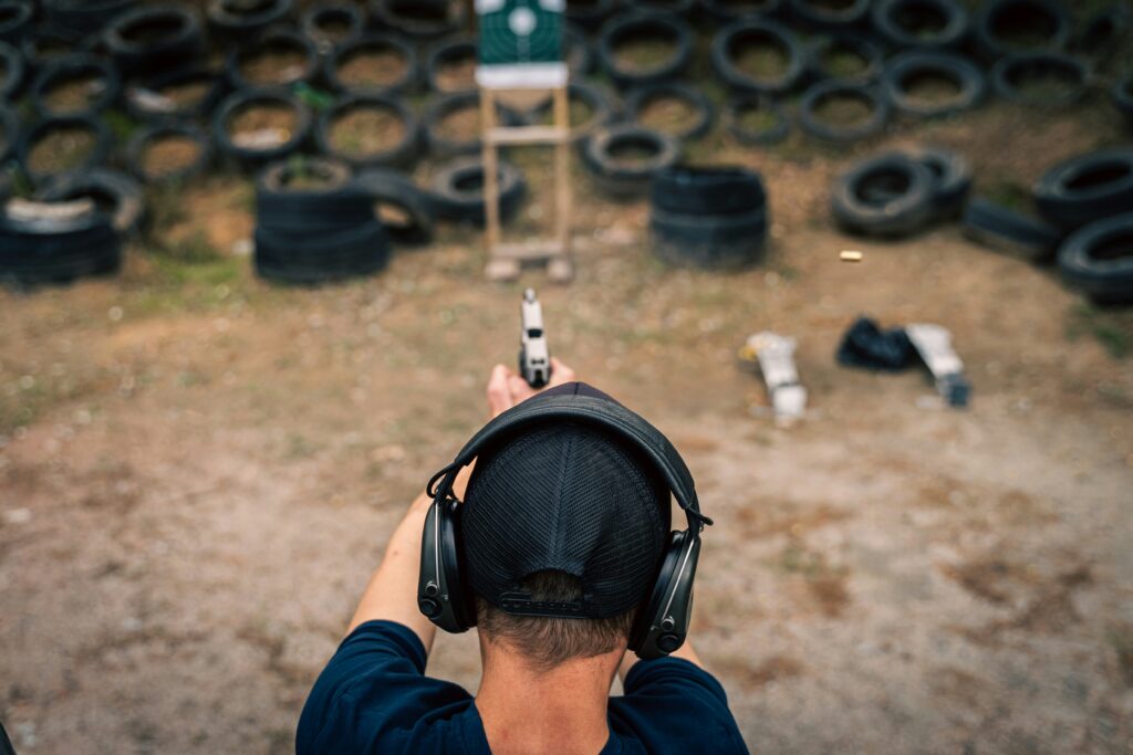 A person wearing ear protection and a cap is holding and aiming a handgun at a shooting range. There are numerous stacked tires in the background and a white target is visible ahead.