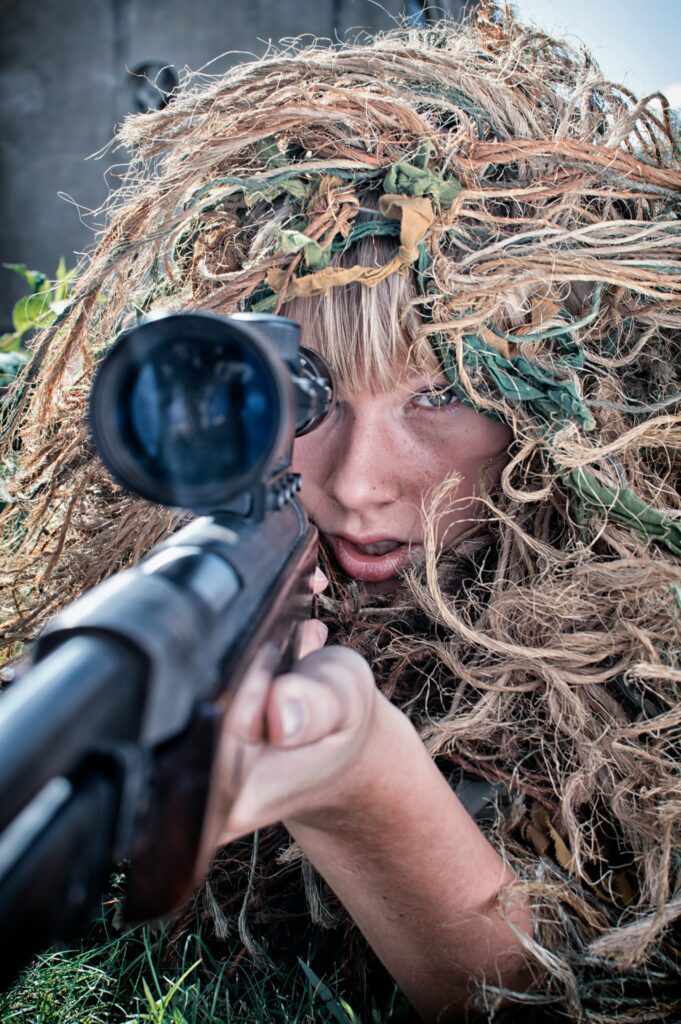 A person in a camouflage ghillie suit aims through the scope of a sniper rifle, lying prone on the grass. The suit is covered in natural foliage, blending with the surroundings. The focus is on the intense gaze through the scope.