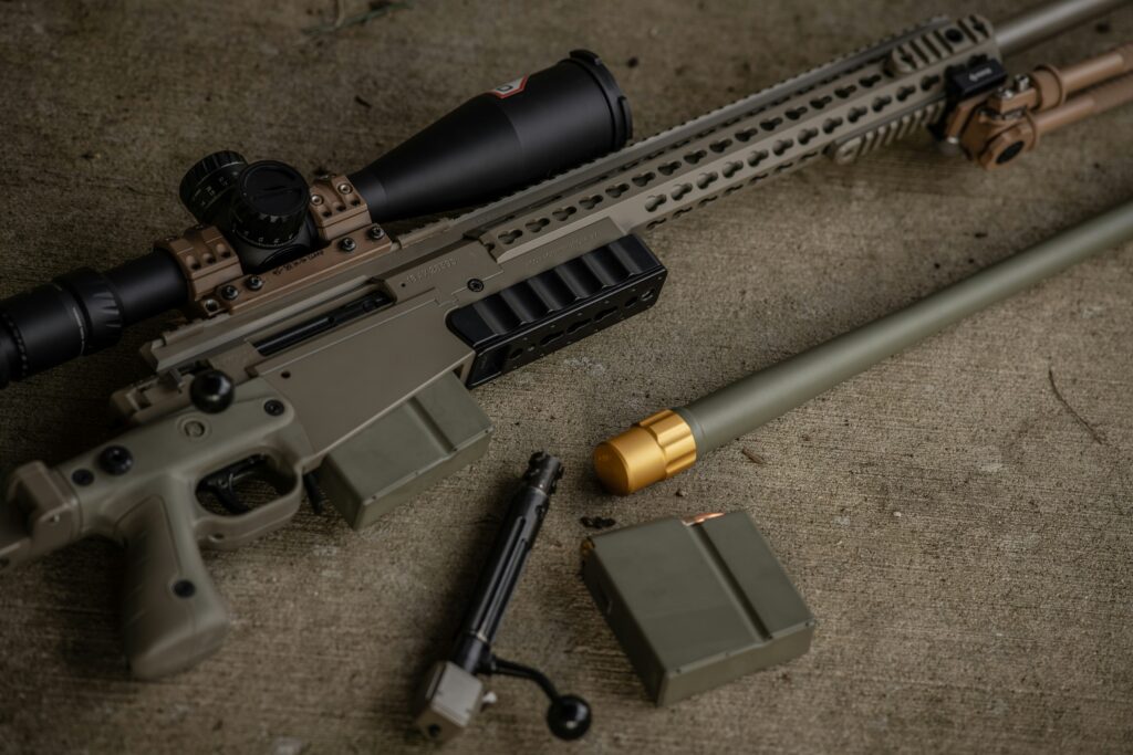 A disassembled firearm lies on a gray surface. Components include a rifle with a mounted scope, a bolt, green magazine, a green barrel with a gold tip, and various small gun parts. The color scheme is mostly green and matte black.