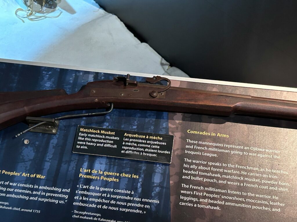 A display of a historical musket atop an informational panel. The musket has a wooden stock and metal parts, typical of the period. The panel beneath provides details about Comrades in Arms, contrasting French and Indigenous warriors.