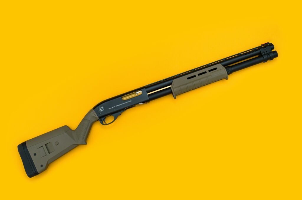 A shotgun with a matte finish displayed diagonally on a vibrant yellow background. The stock and grip are olive green, while the barrel and other metal parts are black, featuring a gold-tone element near the trigger guard.
