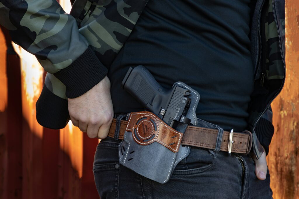 A person in a camouflage jacket is carrying a black handgun in a brown leather holster attached to their belt. The background shows a rusty metal wall.