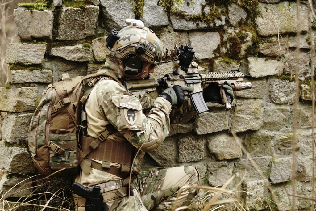 A soldier in camouflage and tactical gear crouches by a stone wall, aiming a rifle. The scene is outdoors, and the wall has patches of moss. The soldier carries a backpack and appears focused on a target.