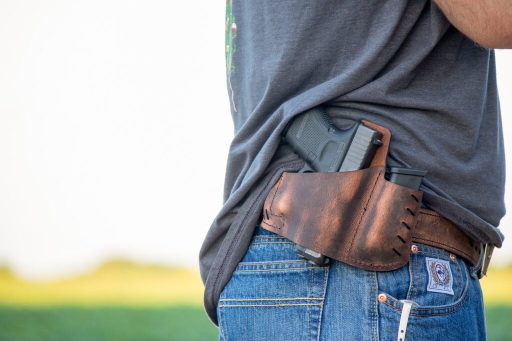 A person wearing jeans and a gray T-shirt has a handgun in a brown leather holster on their waist, partially concealed. A green field and pale sky are in the background.