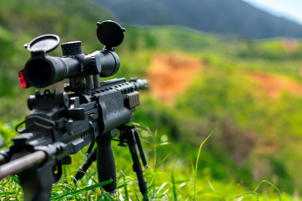 A close-up of a sniper rifle with a scope, mounted on a bipod, set in a grassy field. The background features a blurred view of rolling hills under a clear sky, suggesting a remote, outdoor location.