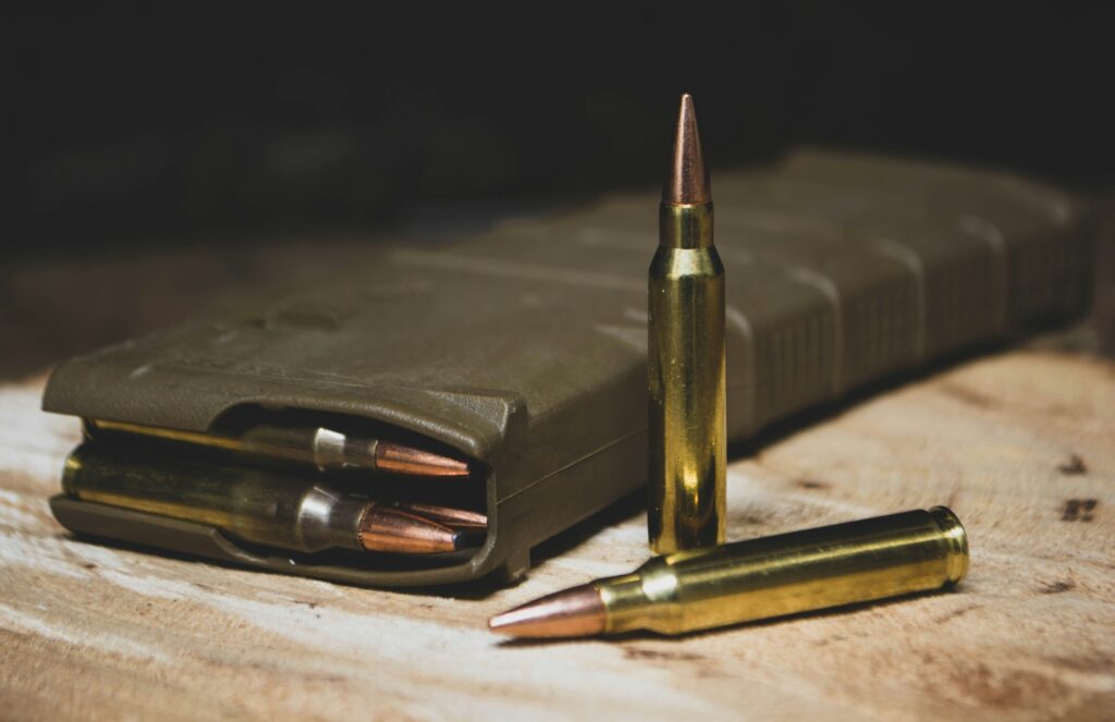 Close-up of a firearm magazine lying on a wooden surface, partially filled with several rifle cartridges. Two bullets are positioned upright next to the magazine, showcasing their shiny brass casings and pointed tips.